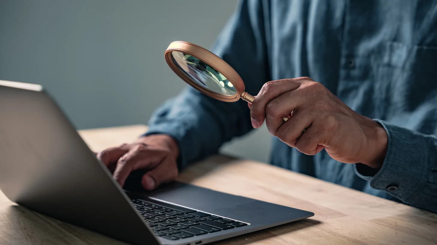 A person holding a magnifying glass beside a laptop while typing on it